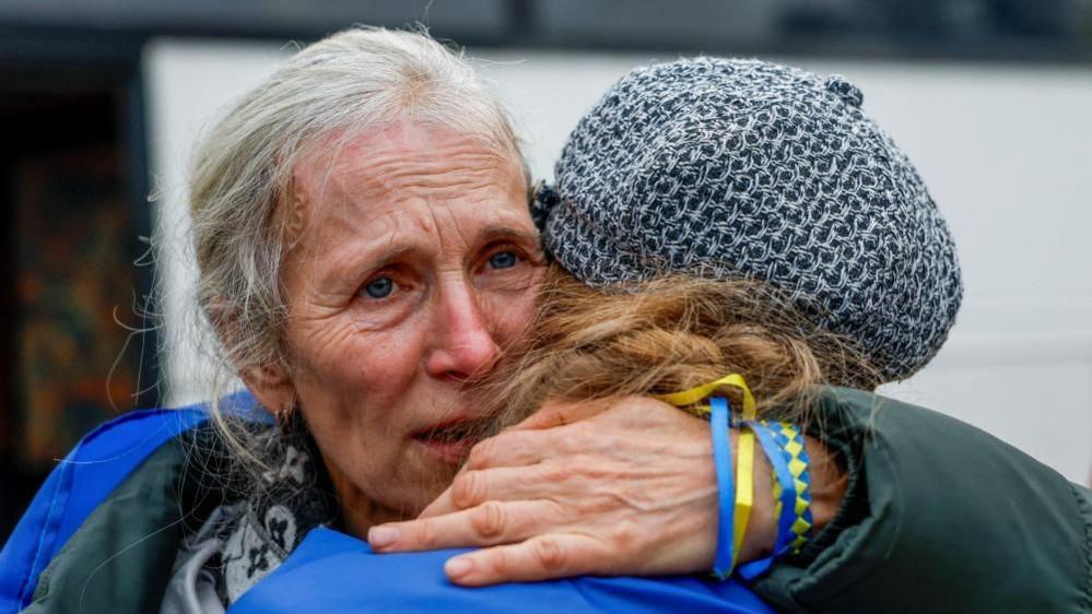 A woman embraces her daughter. She has tears in her eyes and she is wearing several blue and yellow bracelets.