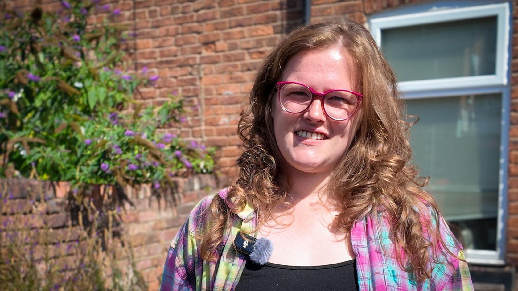 Courtney-Grace is standing outside by a brick garden wall and is in front of a window. She has pink-rimmed glasses, long light-brown wavy hair and is wearing a brightly coloured checked shirt with a black top underneath