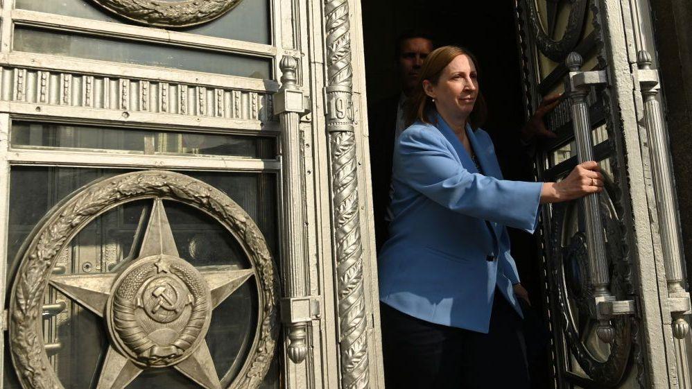 Lynne Tracy walks through a large set of ornate double doors. She has short brown hair and wears a cornflower blue blazer. She is pushing open the left door using a long vertical handle. The doors are engraved with bronze-coloured symbols, including the hammer and sickle.