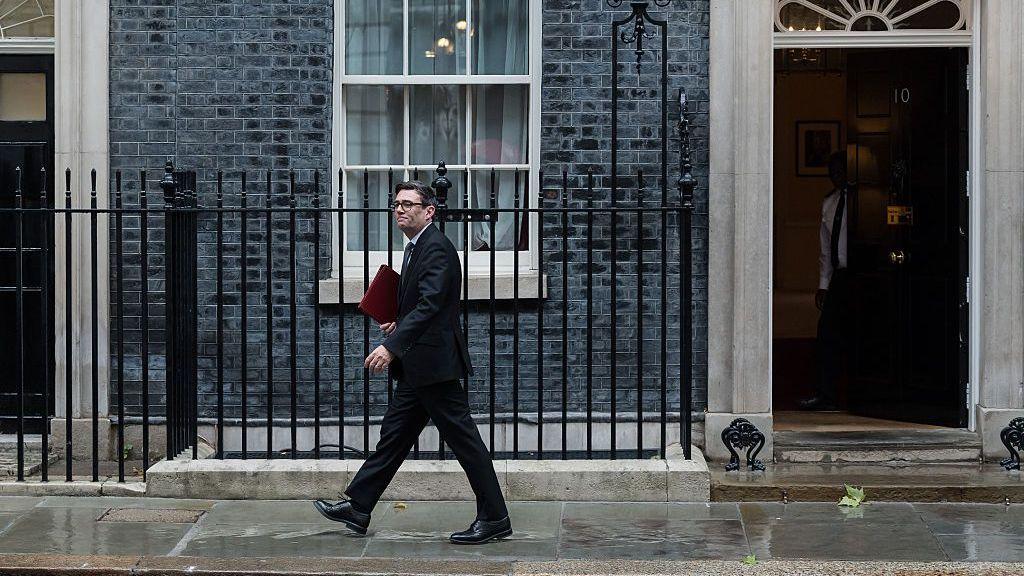 Andy Burnham walks along the pavement in Downing Street in front of the exterior wall and the black Number 10 door.