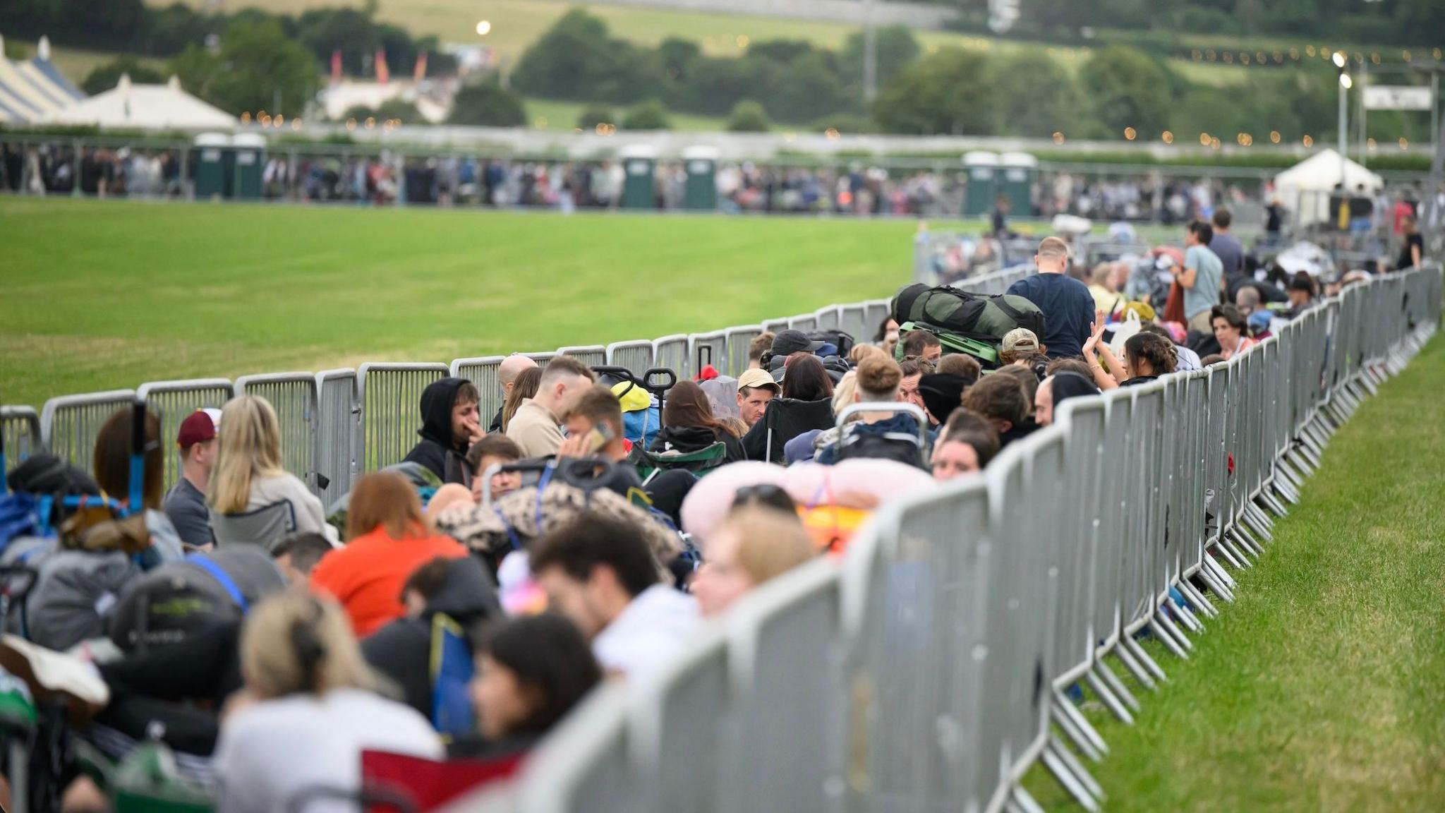 A busy queue of people waiting to get into Glastonbury Festival. They are sat and stood inbetween silver gates in a field.