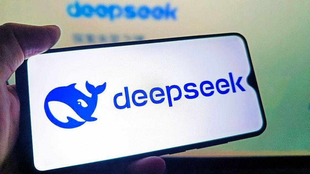 A hand holds a mobile phone, displaying the Blue deepseek logo of a blue cartoonish whale on a white background.