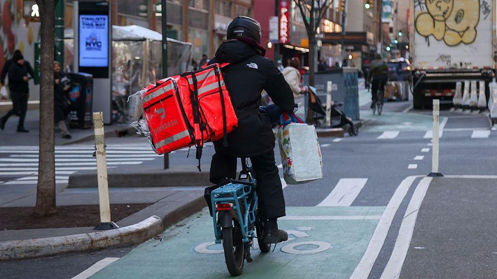 A delivery worker dressed in black rides a bike with a DoorDash bag in New York.