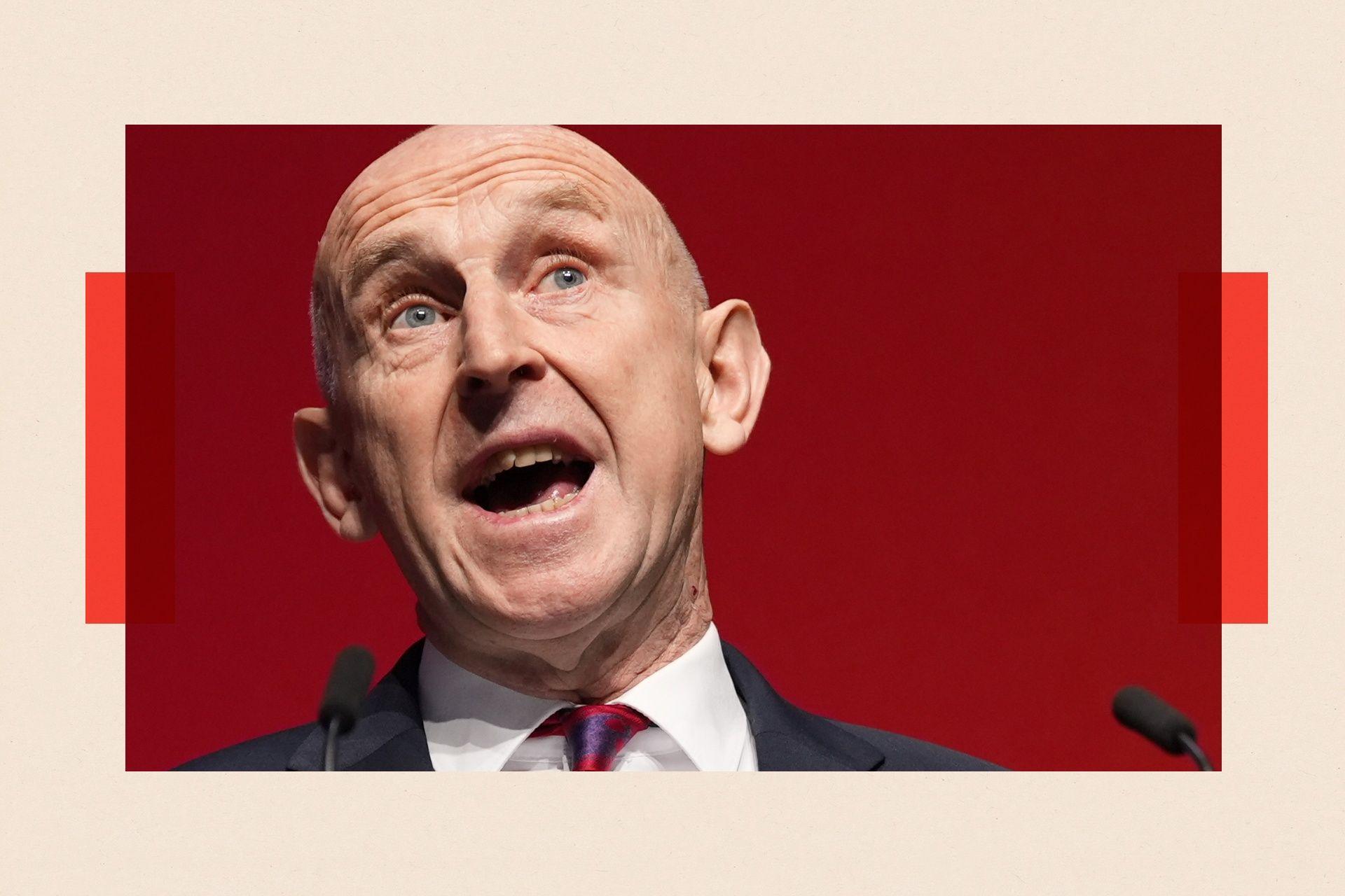 John Healey wearing a white shirt with a red and purple tie, speaking at a podium into microphones.
