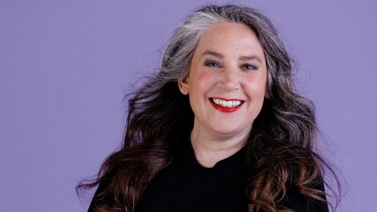 A woman with brown hair that has highlights of grey at the front smiling. She is wearing dark eyeshadow and red lipstick, and is wearing a black jumper. The background is purple.