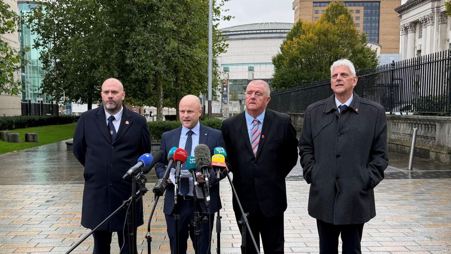 Four men stand behind six microphones. They stand on light-grey and beige bricks in front of white-stone buildings.