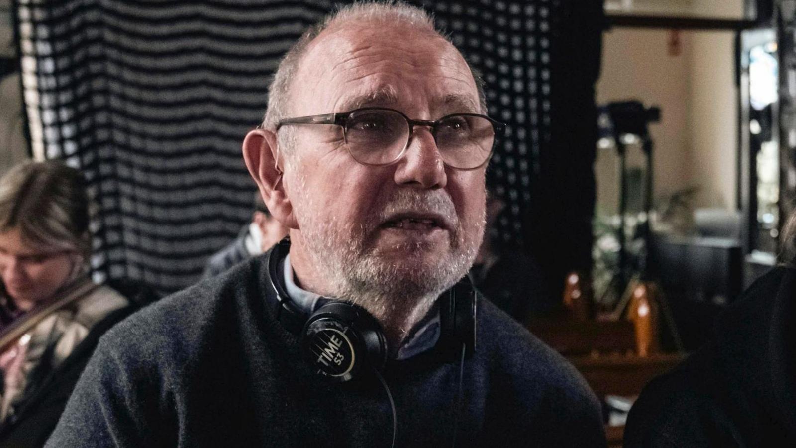 Jimmy McGovern, with short cropped hair, a stubbly beard and glasses, has headphones round his neck with the word Time printed on them. He looks past the camera, while the production team for the BBC drama work in the background