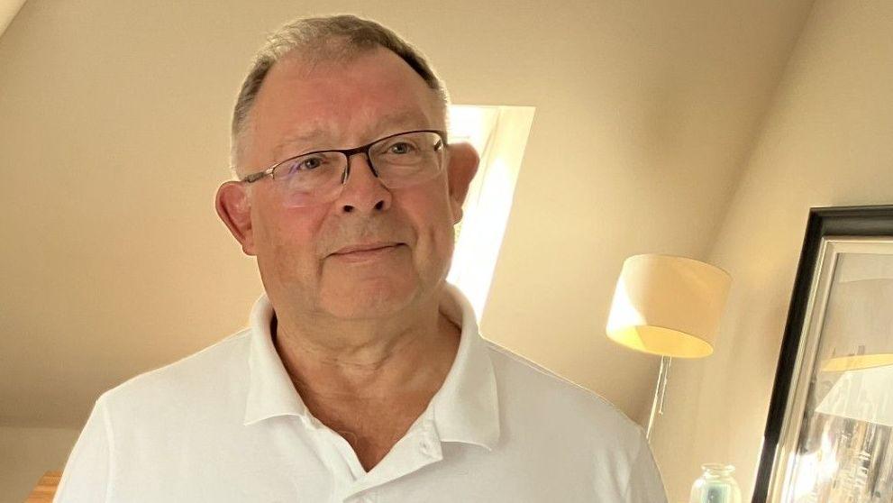 Rod Stone, who is wearing glasses and a white polo shirt, is standing in his living room and is smiling at the camera.