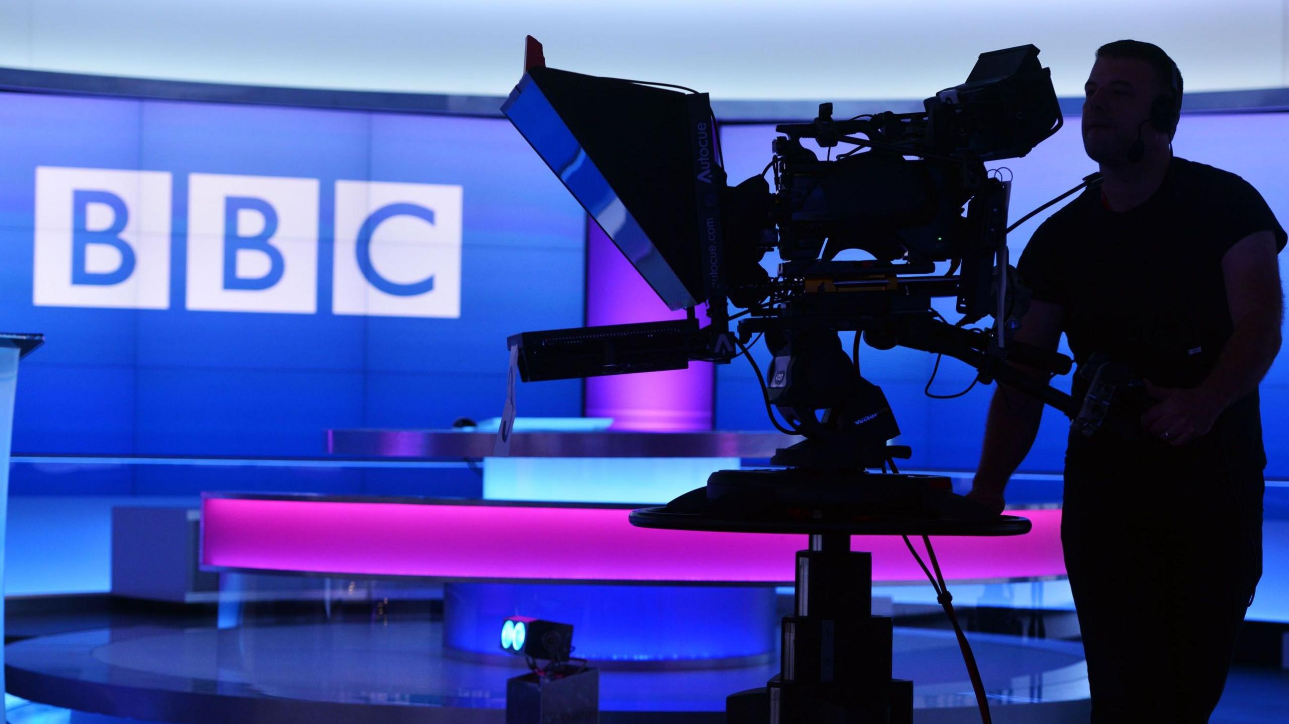 A camera man seen in a studio with the BBC logo in the background against a pink and purple backdrop