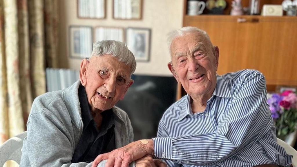 Ronald Hendrey, in a grey fleece over a dark top, is clasping hands with Henry Rice, in a blue and white striped shirt. They are both seated and smiling.