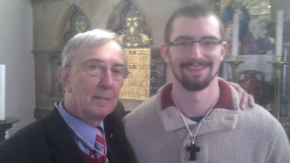 An older man, Peter Farquhar, has his arm around Ben Field who is wearing a crucifix. They are stood in a church.