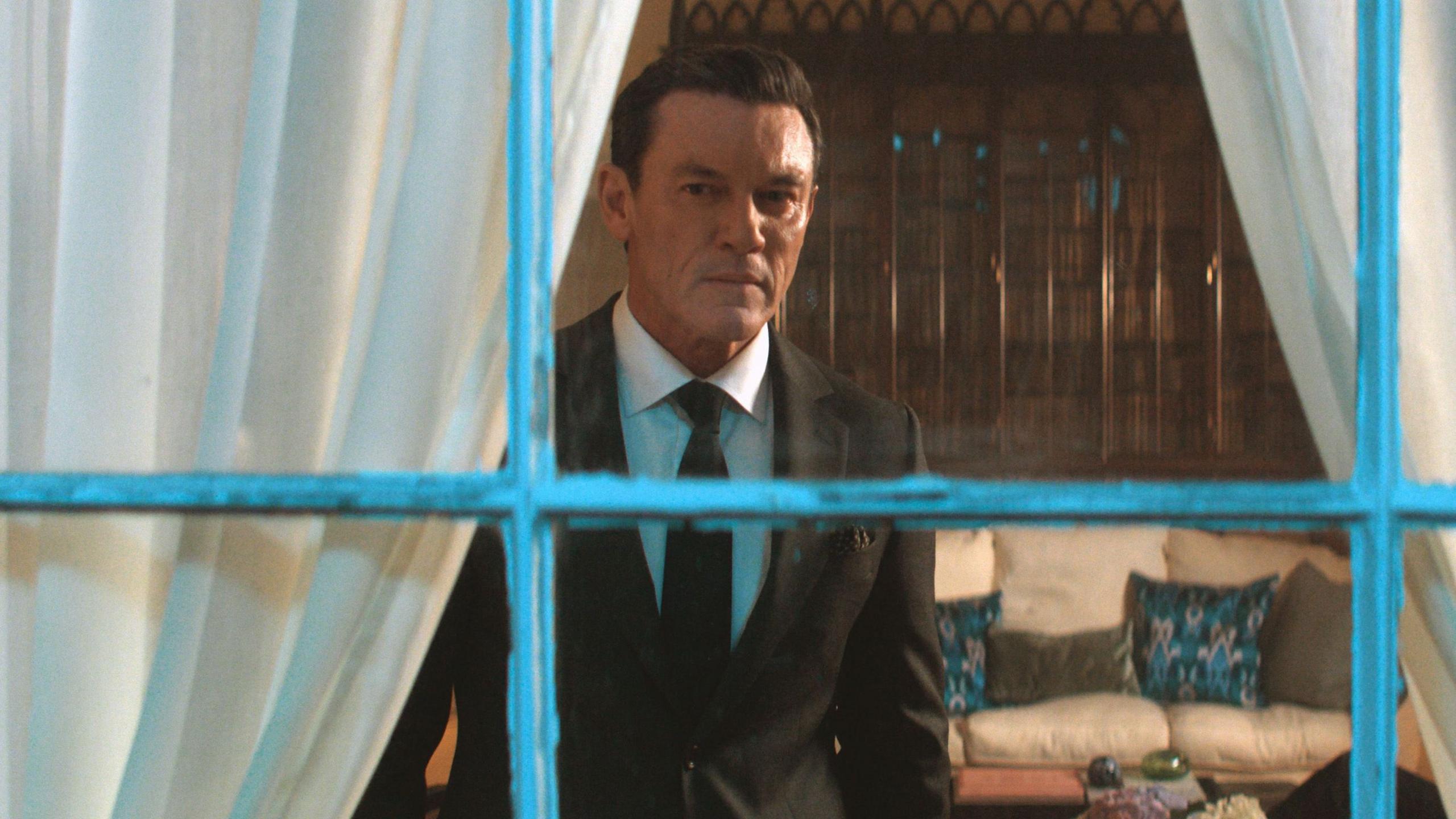 Evans is photographed in character as Martin Gilmour. He is staring past the camera through a window with a chipped white frame. White curtains frame the window from the inside. A white sofa can be seen behind him, with four  cushions on top. He wears a black suit and tie, with a white shirt.