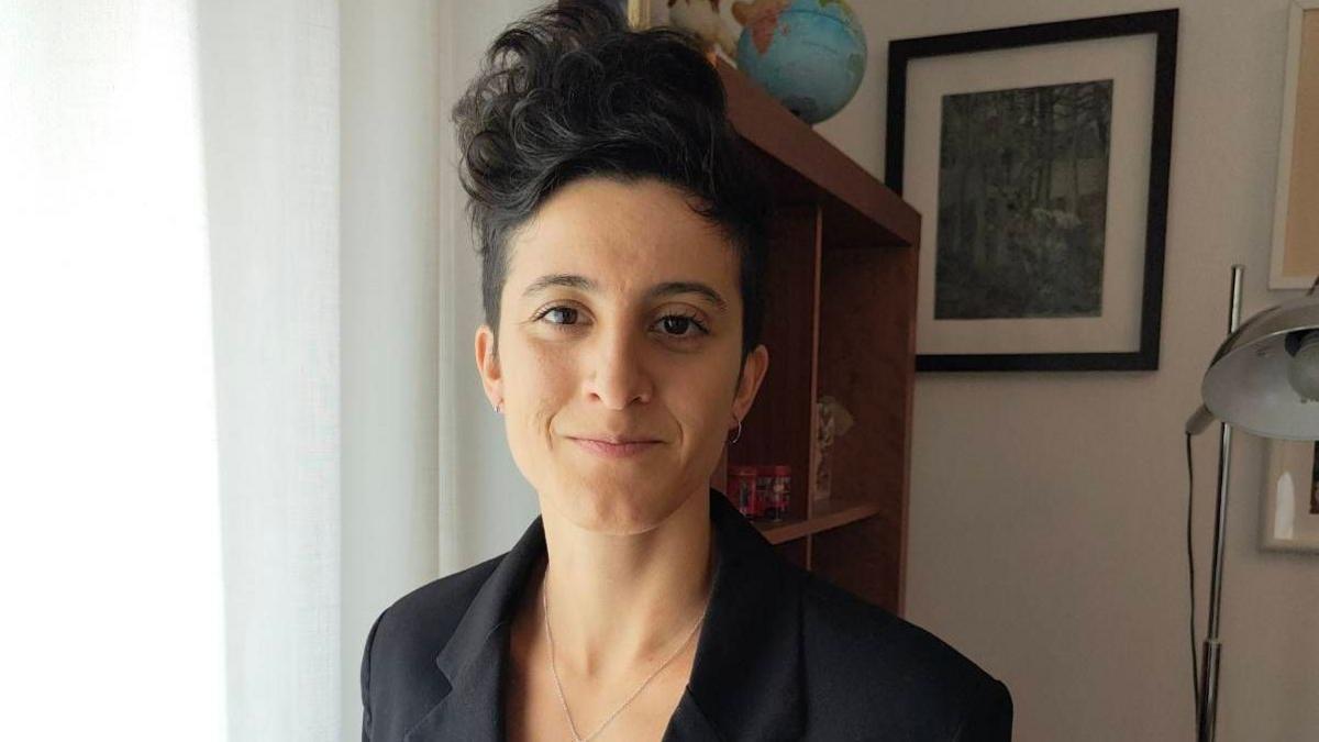 Sofia has short dark hair that is in a quiff style. She is wearing a black blazer and is standing in a room with a lamp and bookshelf in the background.