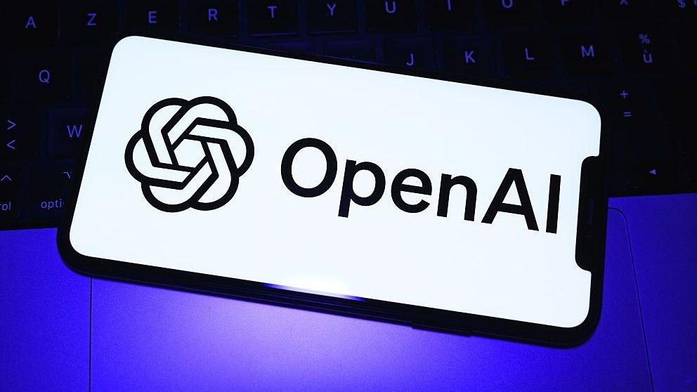 The OpenAI logo in black font on a white phone background over a purple light and black keyboard