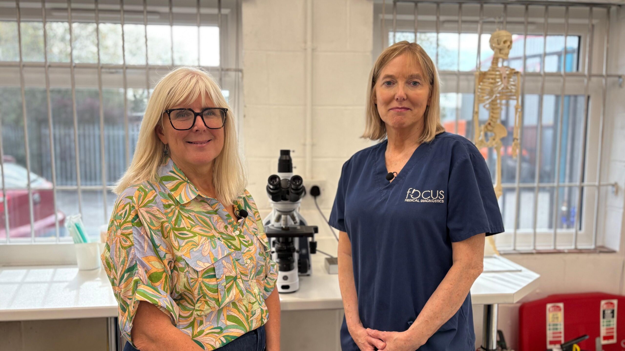 Two women standing either side of a microscope in a laboratory smiling at the camera. The woman on the left has shoulder length blonde hair and is wearing a multi coloured blouse. She is also wearing glasses. The woman on the right is wearing a blue medical uniform with Focus Medical Diagnostics in white writing on her top.