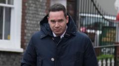 A man with short black hair wearing a large dark blue coat. He is looking into and walking towards the camera
