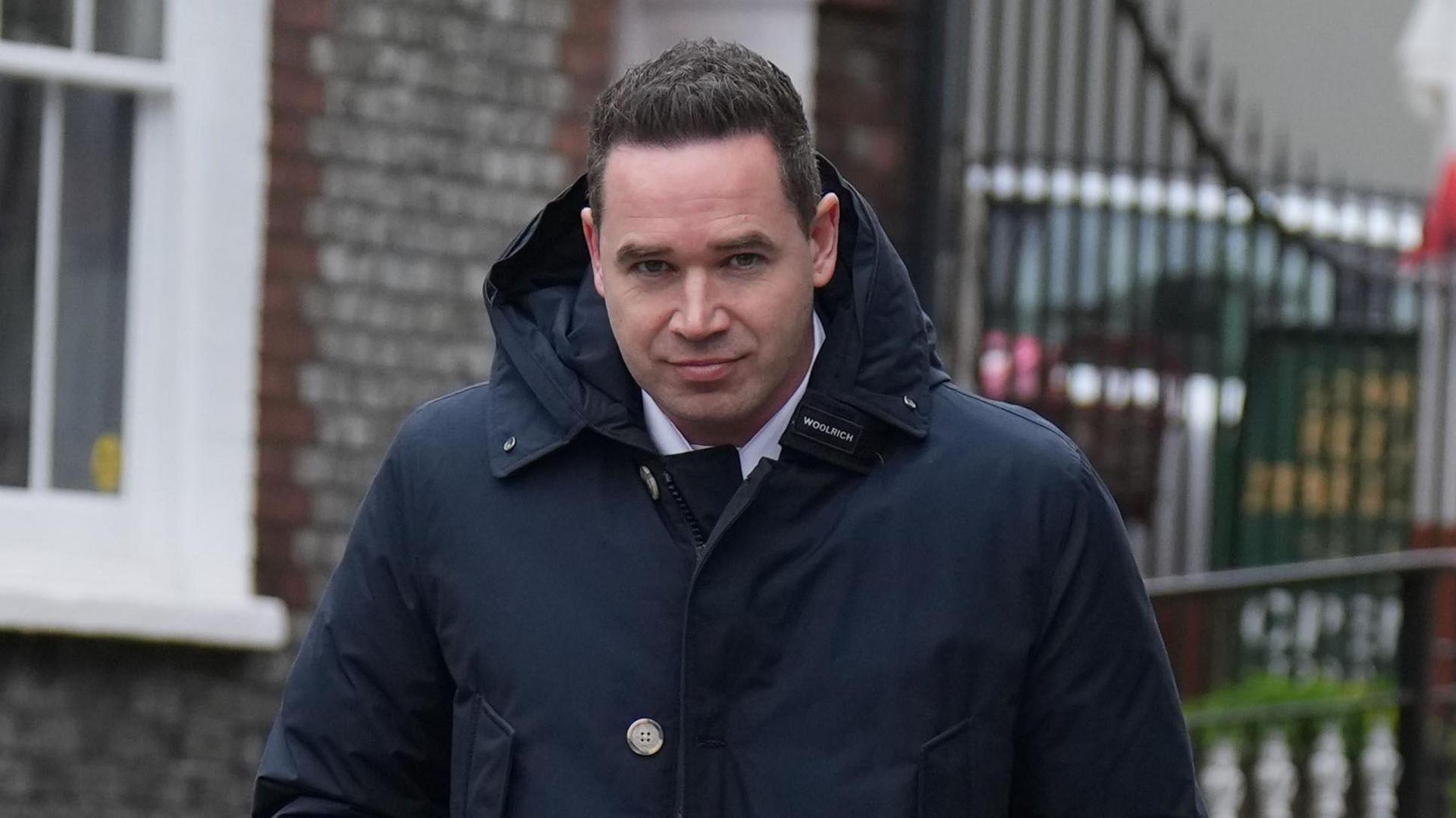 A man with short black hair wearing a large dark blue coat. He is looking into and walking towards the camera