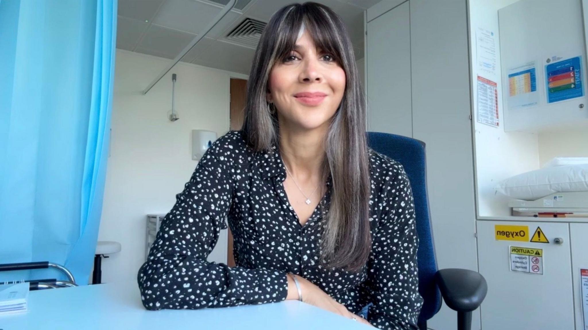 A woman with long brown hair and a fringe smiling, she is sat down on a blue chair and is wearing a black blouse with white polka dots, the background is  doctor's clinic.