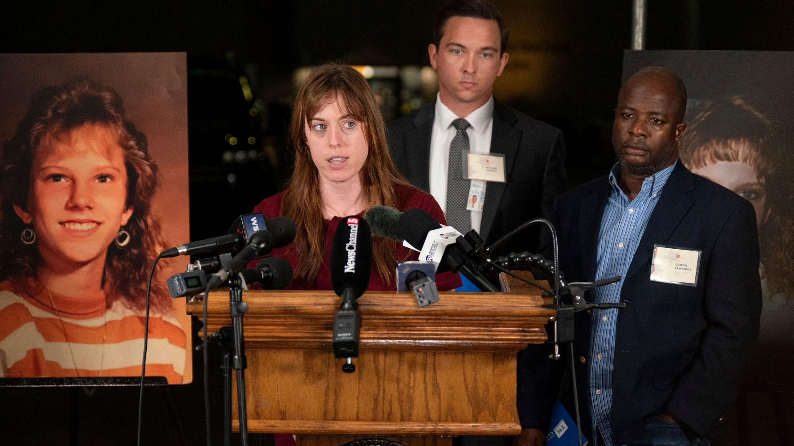 A woman with lonb brown hair flowing over a burgundy blouse stands in front of a podium outfitted with multiple microphones, two suited men standing behind her, as they are flanked with enlarged photos of a blonde and smiling teenager, Colleen Slemmer, who was murdered at age 19 by Christa Pike