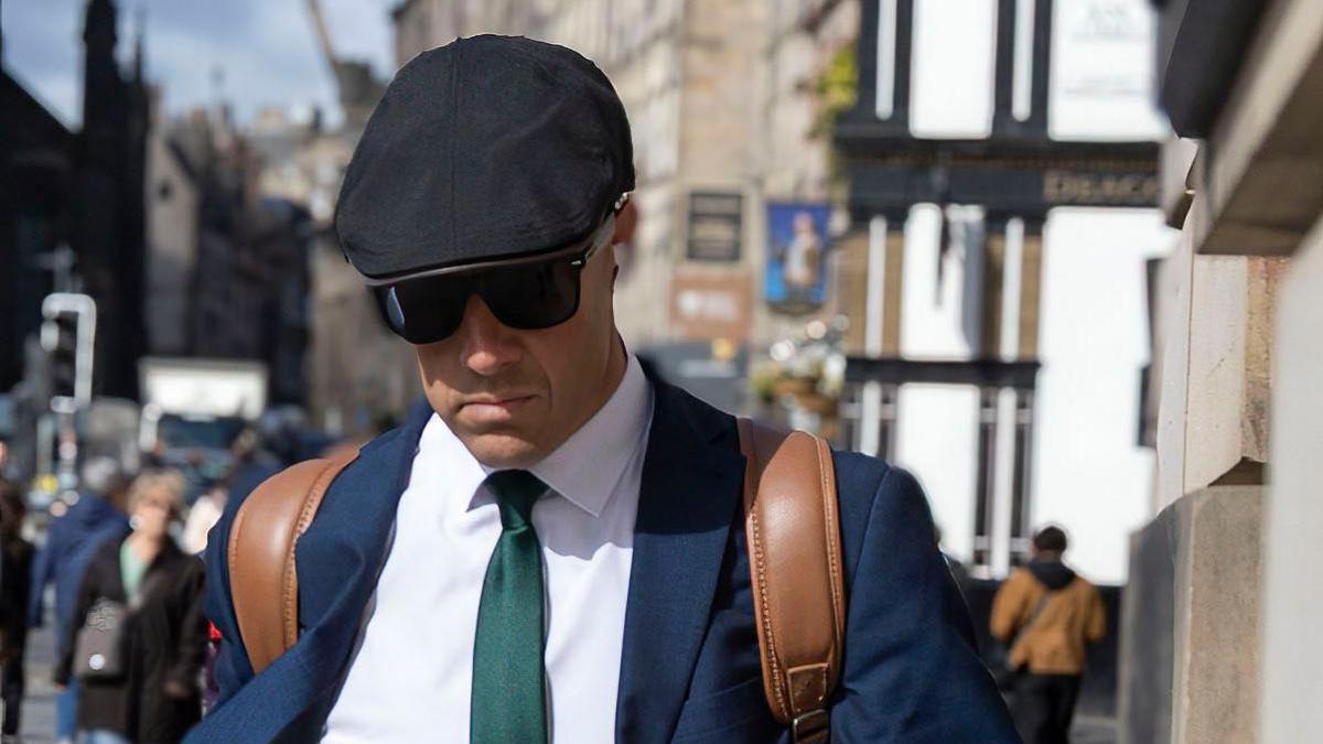 A man keeps his head down as he walks along a street. He is wearing a black flat cap, sunglasses, a blue jacket, white shirt, green tie and has leather rucksack handles over his shoulders
