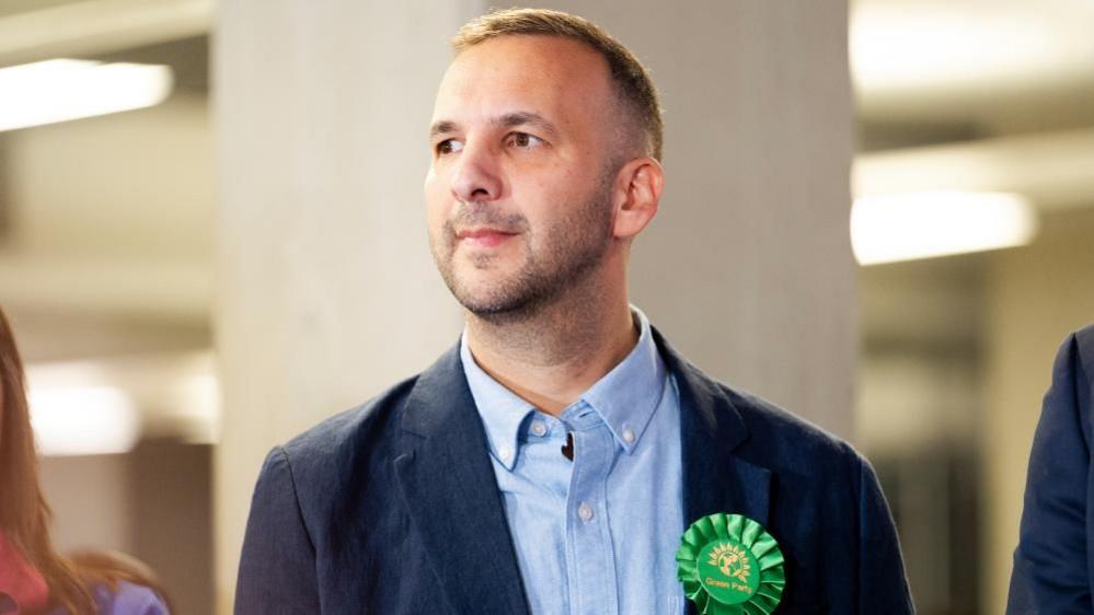 Zack Polanski looks to his right while on stage at the by-election count where the results were announced. He is wearing a dark blue suit, light blue shirt and his green rosette.