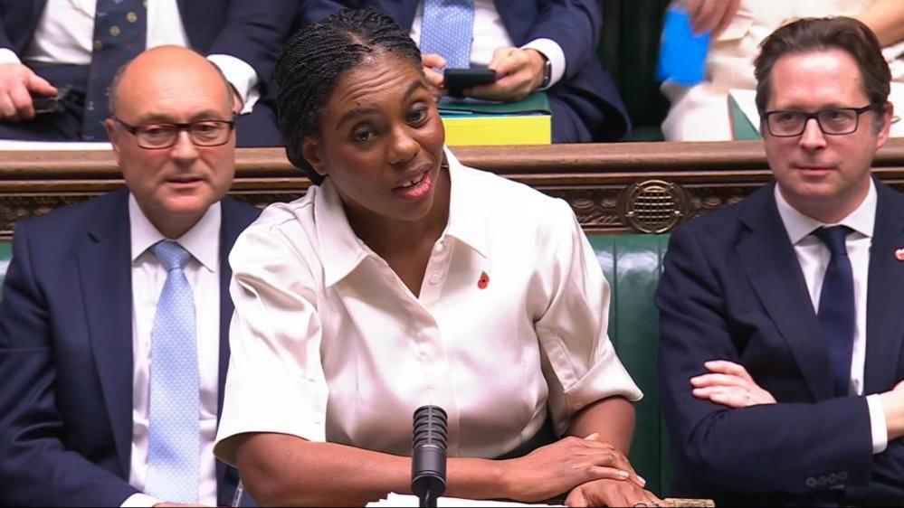 Kemi Badenoch leans on the dispatch box, wearing a cream short-sleeved blouse with a poppy pin on her left side, flanked by two shadow ministers both wearing navy suits with white shorts