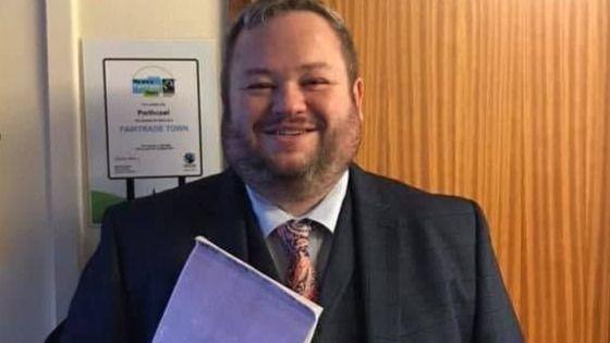 Gareth Ritter pictured smiling and wearing a dark suit and a matching waistcoat and patterned tie holding a purple exercise book. He has short brown hair and facial hair.