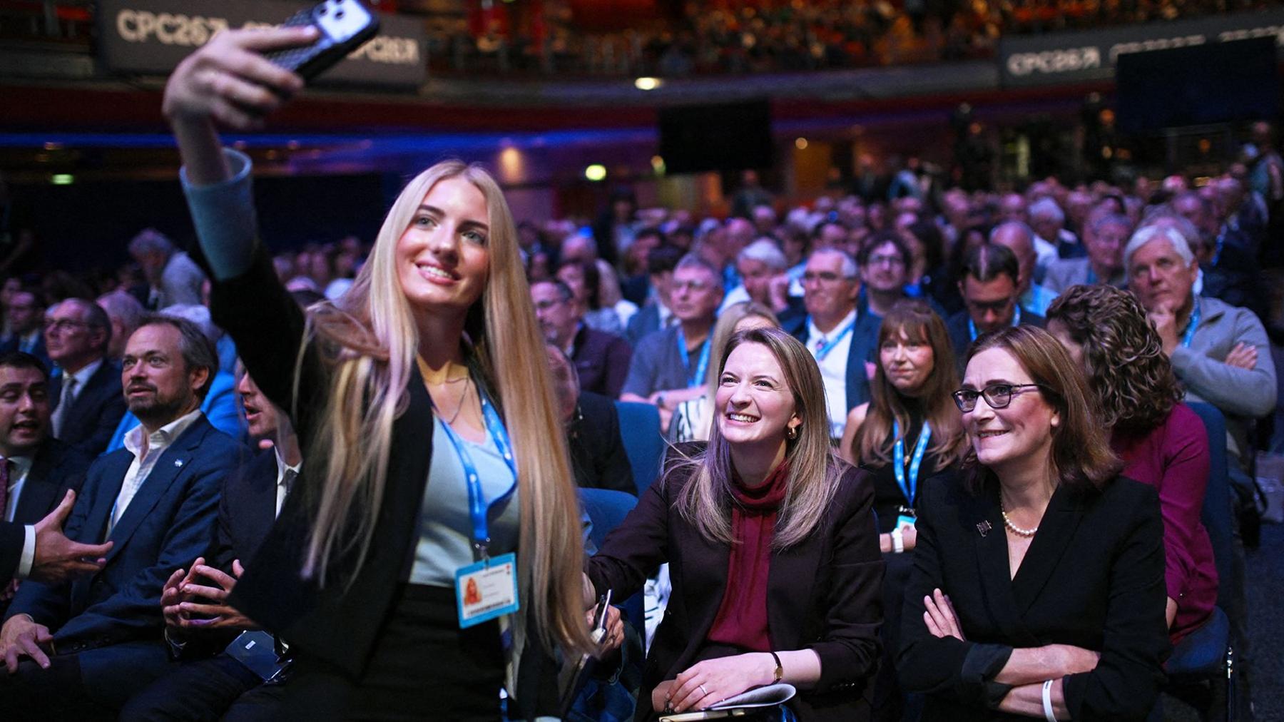 A young woman with long blonde hair takes a selfie with shadow housing secretary Katie Lam, who is sitting in the conference hall with Tory members