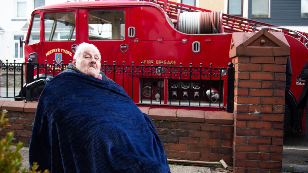 Dave Evans, wrapped up warm in a blue blanket, sits in front of a vintage red fire engine