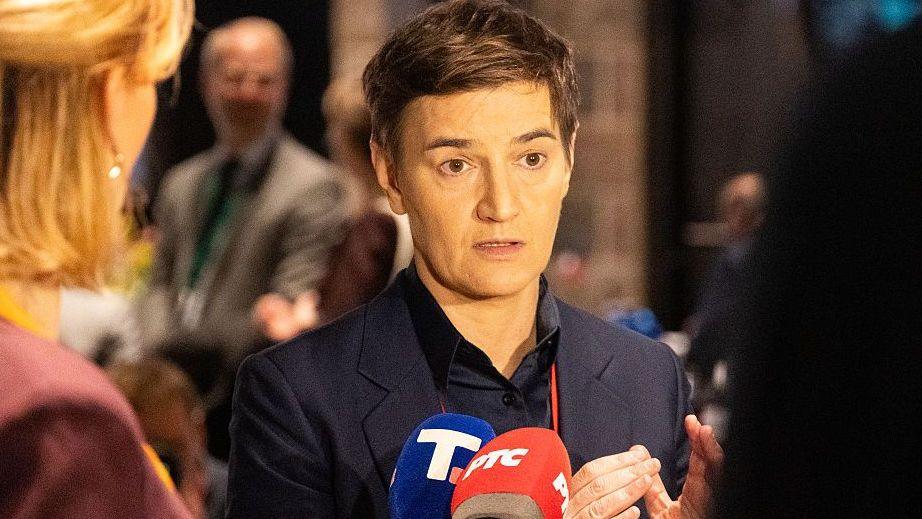 A woman with short hair and a jacket talks to reporters