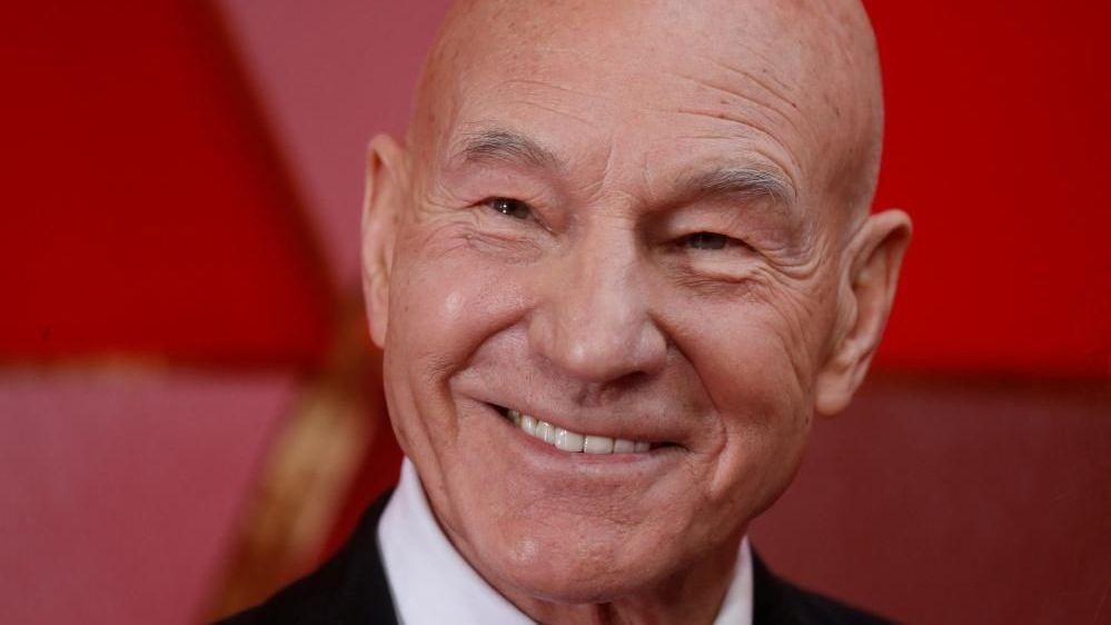 Sir Patrick Stewart smiling, in front of a red background