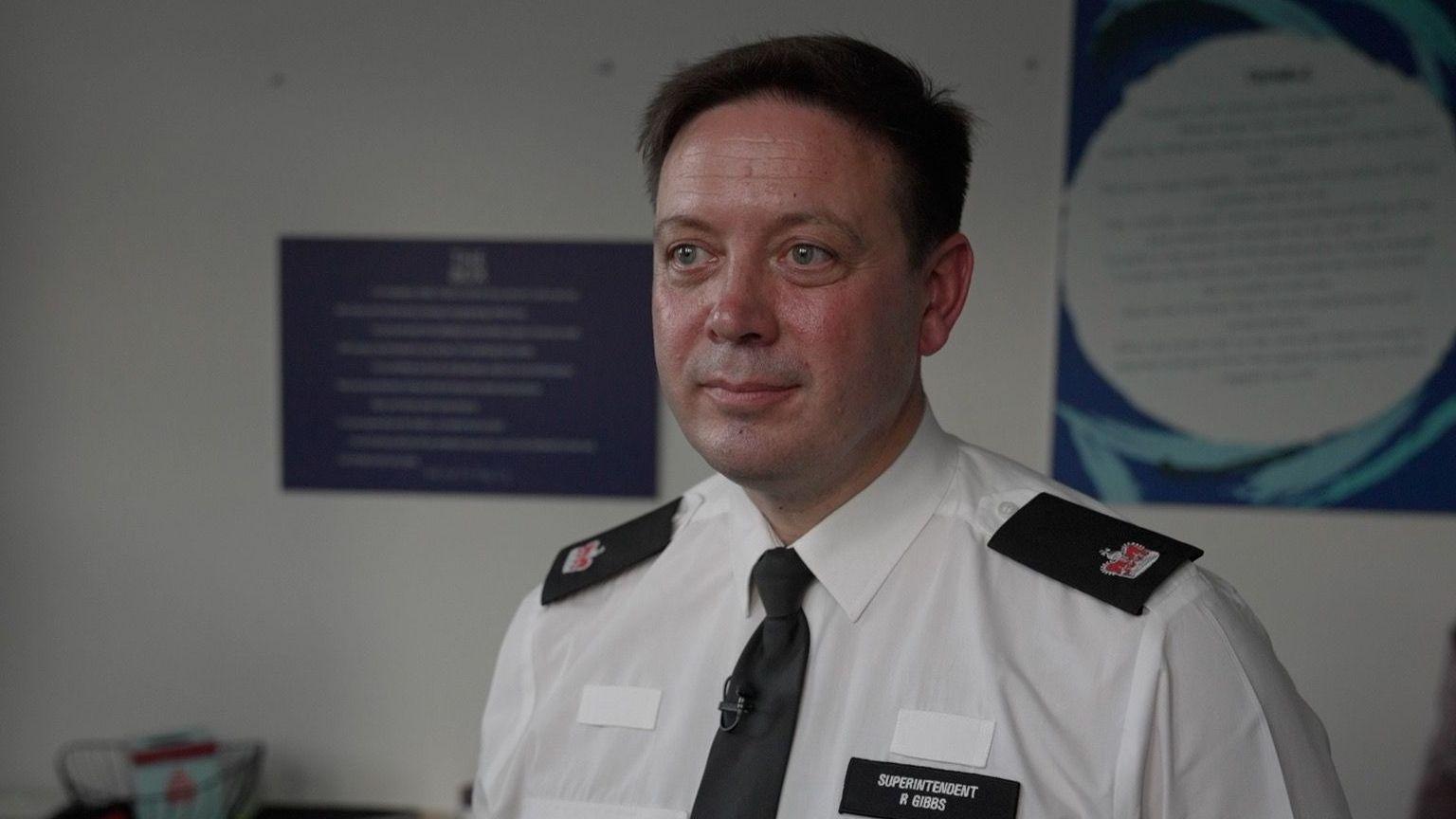 Supt Rob Gibbs in uniform stands with his back to the wall and looks away from the camera