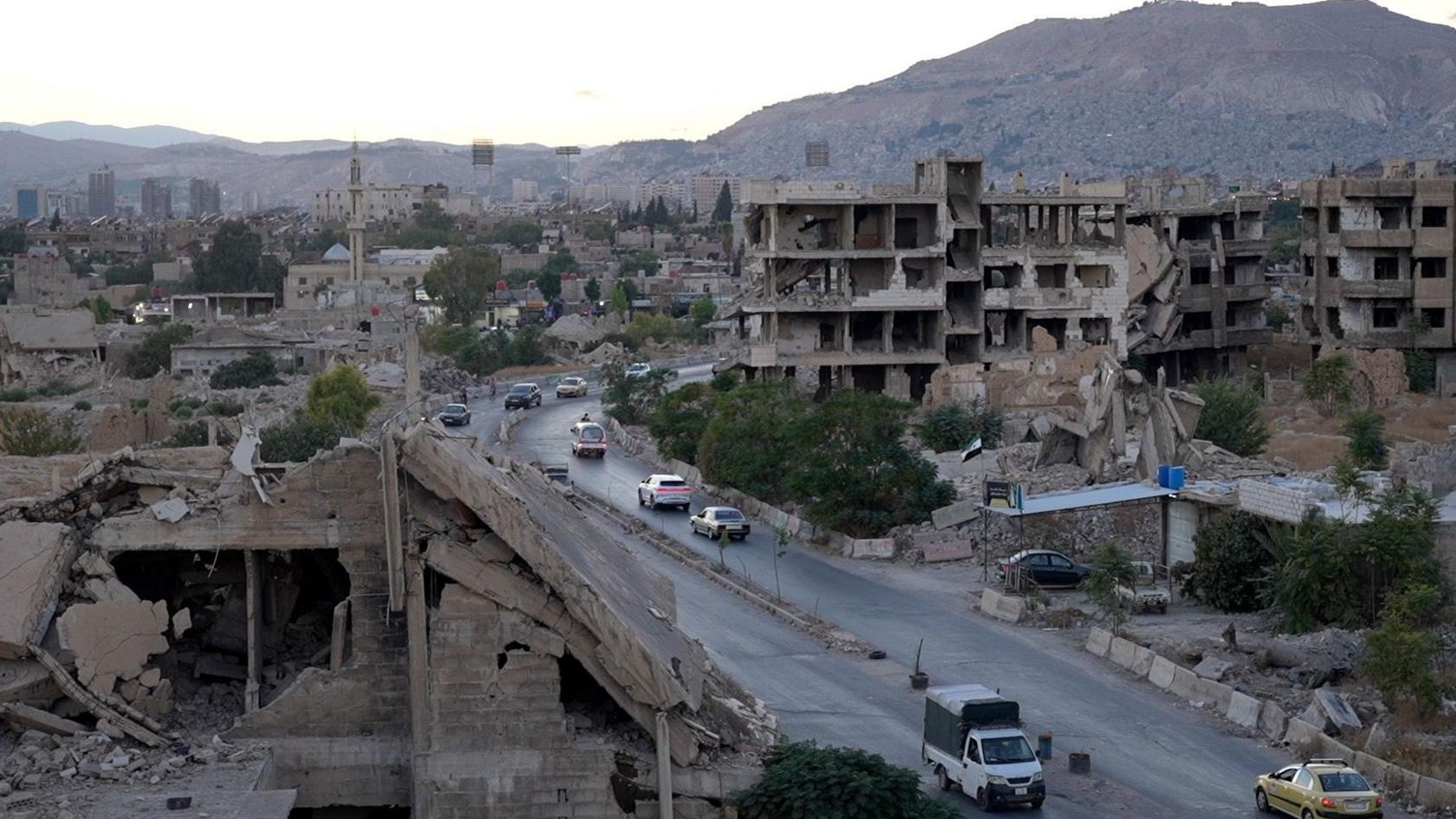 Wide shot shows a built up urban area with several tall buildings seriously damaged, a number of cars driving on a road running through the centre and mountains in the background
