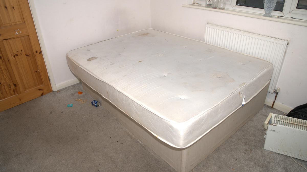 A dirty white mattress is pictured with no bed sheets on, and various items on the floor.