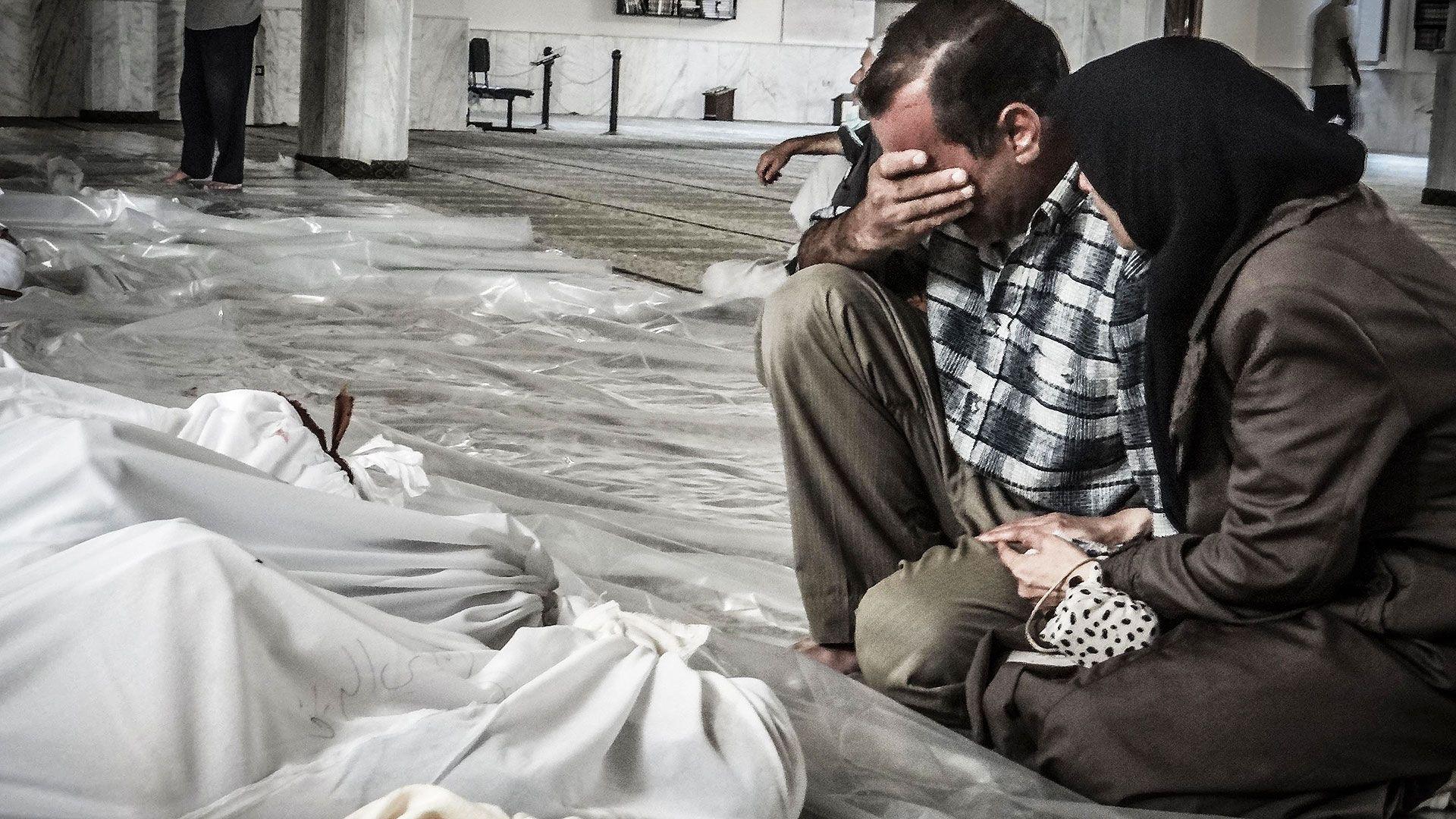 A mother and father weep over their child's body killed in a suspected chemical weapons attack on the Damascus suburb of Ghouta, in August 2013. The man is covering his eyes, wearing a black and white shirt and khaki coloured trousers. The woman is wearing a brown coat and black hijab. They are sitting next to bodies wrapped in white sheets.