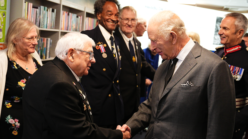 King Charles is wearing a grey suit and tie, shaking the hand of a man wearing a black suit and medals.