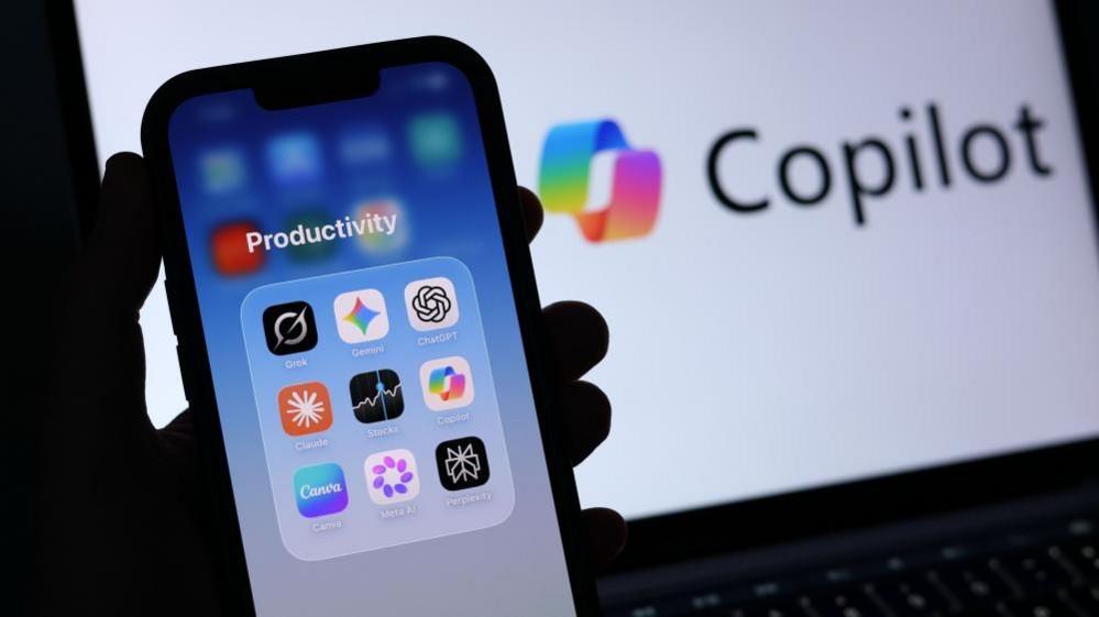 A silhoutted hand holds an iphone, showing ann app folder containing open AI products. Behind it, a laptop screen shows the loho for Microsoft Co-pilot.