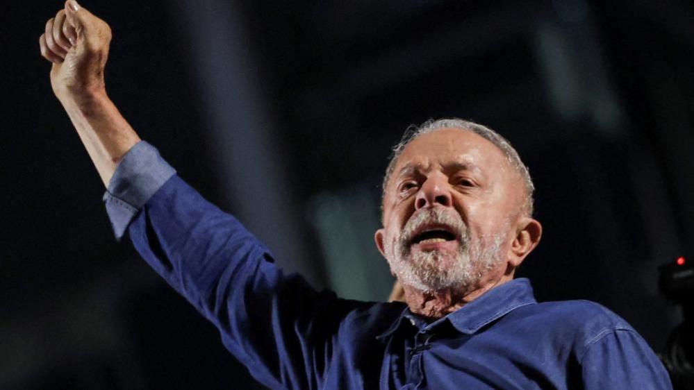President Lula extends his right arm, his hand in a fist, as he reacts to his election. He is wearing a blue denim shirt, and has short grey hair and beard.