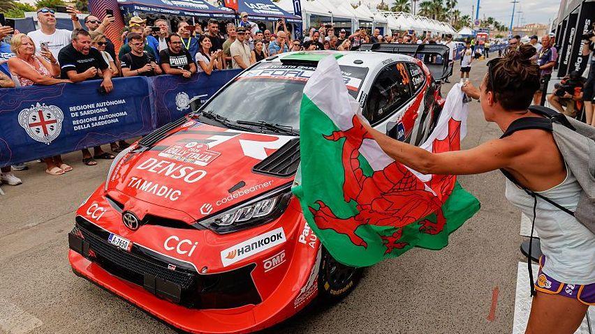 A suporter holds a Welsh flag in front of a rally car as crowds behind cheer