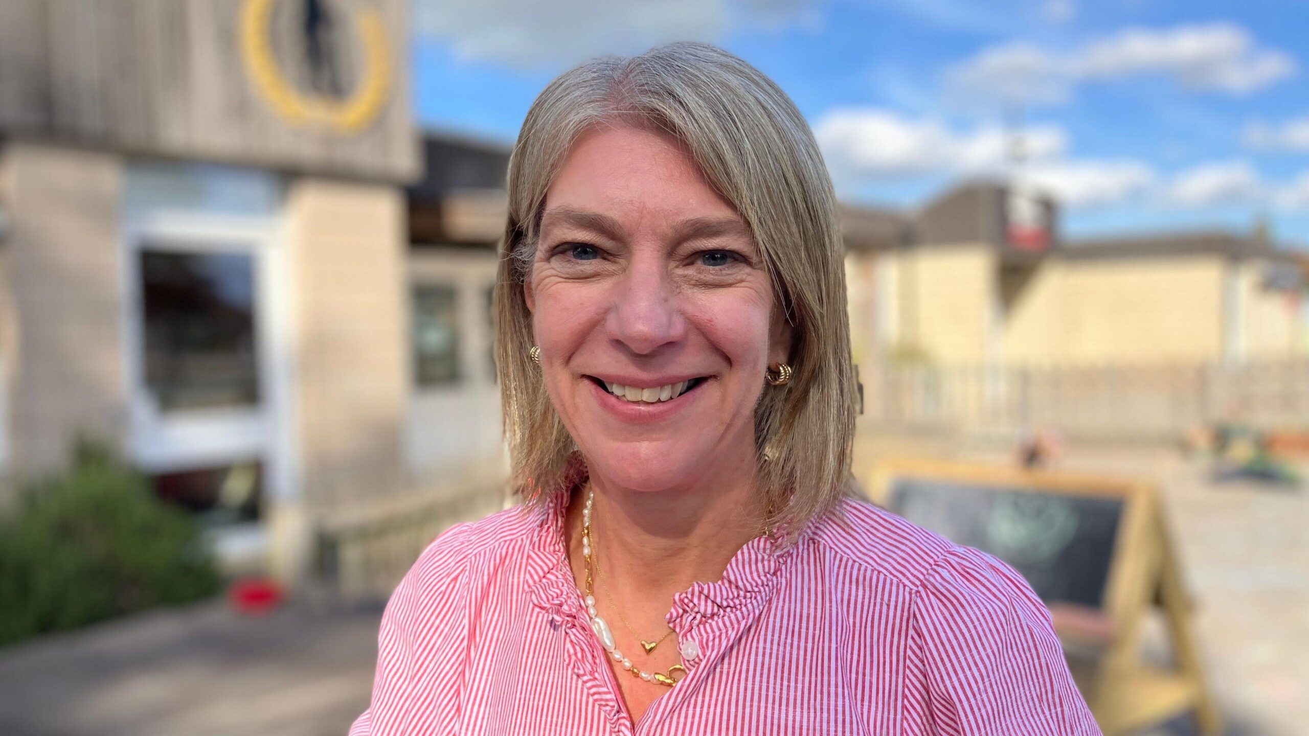 A close-up portrait of Jo Mould, who has shoulder-length grey hair and is wearing a pink striped blouse and gold jewellery, standing outdoors. Buildings and a fenced play area are blurred in the background under a blue sky with scattered clouds.