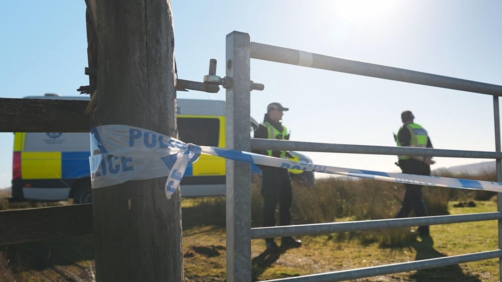 Police cordon in a field with officers and a car parked by a gate on a sunny day