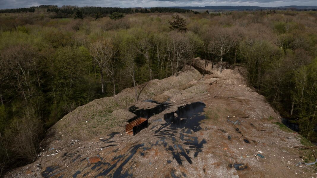 £15m spent cleaning illegal waste supersite in ancient woodland An aerial view shows a woodland clearing that is covered by dumped waste. The rubbish is piled high on the edge of the surrounding treeline and amid the waste are pools of dark water and an abandoned container. The trees stretch to the hills on the horizon, underneath a cloudy sky.