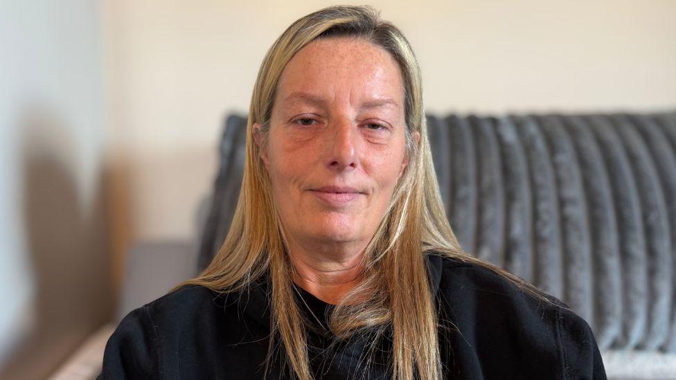 Jill kenning, who has long, blonde hair, wears a black sweater and sits on a grey sofa.