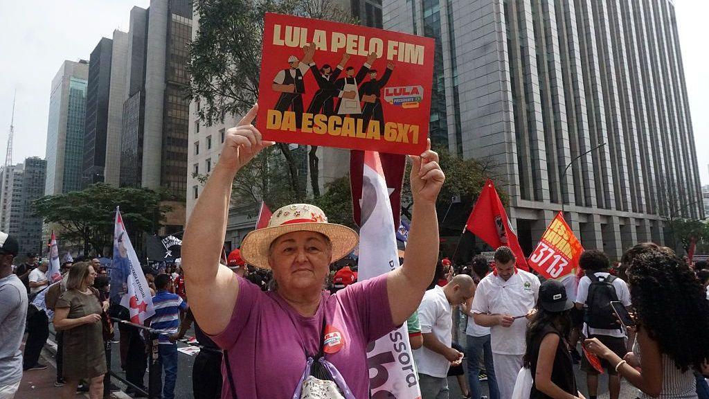 A woman wearing a purple T-shirt and a straw hat holds up a sign reading "Lula for an end of the 6x1 workweek". Behind her other protesters are gathered, taking part in a rally in São Paulo on 30 August, 2026.