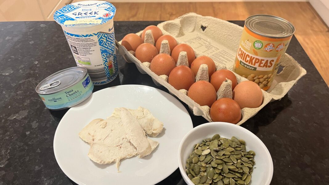 I’m 12 and I want to try protein powder – should I? The photo shows a tin of tuna, a plate of cooked chicken, a bowl of pumpkin seeds, a tub of greek yoghurt, a carton of eggs and a can of chickpeas laid out on a black kitchen work surface. You can see a wooden floor in the background.