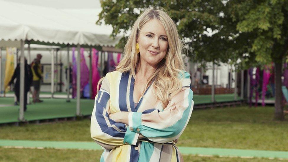 Sophie Howe, a woman with long blonde hair and a green, blue and yellow striped dress, folds her arms while standing in a field with a marquee in the background.