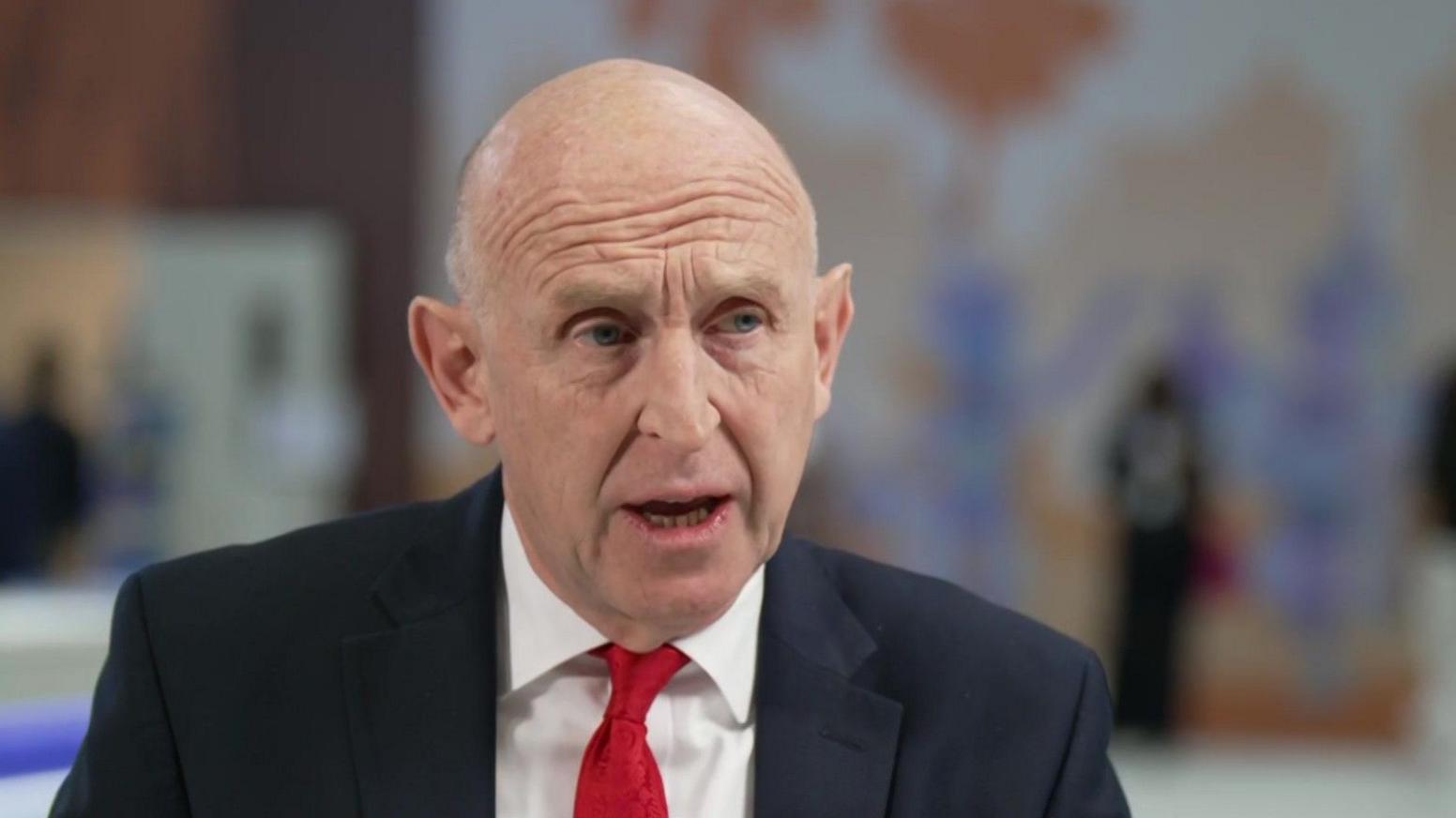 John Healey wearing a dark suit, white shirt and red tie