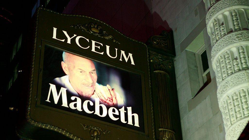 Sir Patrick's face seen on a screen advertising Macbeth outside a theater