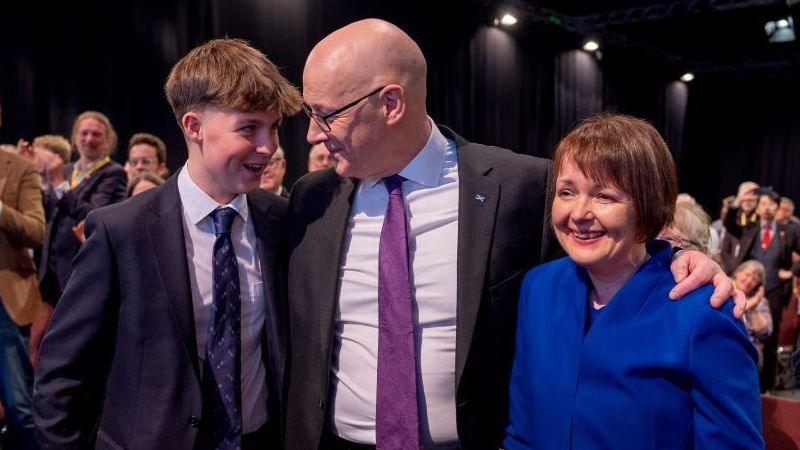 John Swinney, a bald man with glasses wearing a suit, pictured with his son Matthew - a young man in a suit smiling at his dad, and wife, Elizabeth Quigley, at the SNP conference