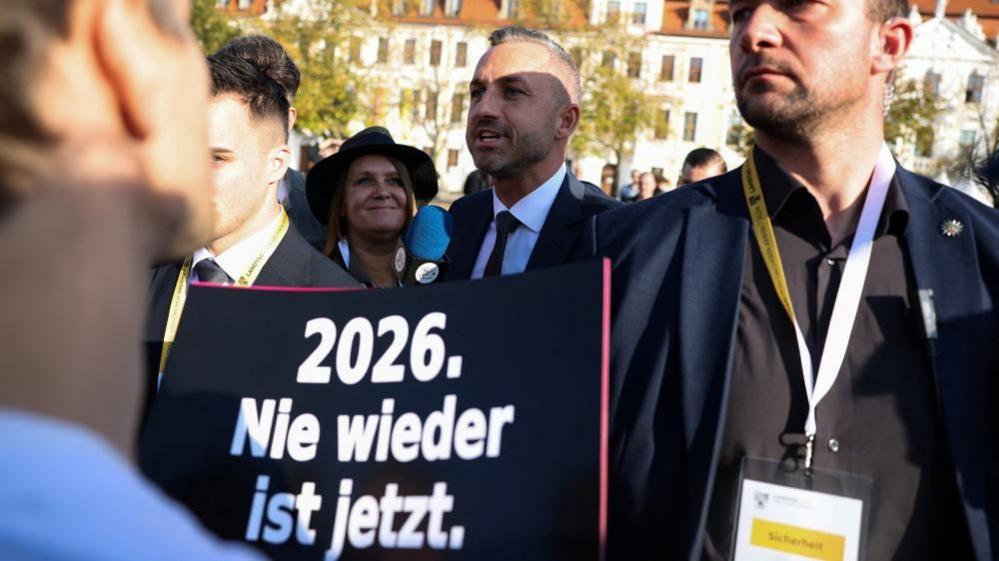 Alternative for Germany's (AfD) Ulrich Siegmund speaks to the media as protesters demonstrate against the election of an AfD candidate as new president of the state parliament of Saxony-Anhalt, in Magdeburg, Germany, October 6, 2026. The sign reads: "2026. Never again is now."